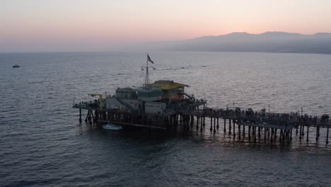 Toma-Panorámica-Aérea-De-Primer-Plano-Del-Restaurante-Al-Final-Del-Muelle-De-Santa-Mónica-En-Una-Concurrida-Noche-De-Verano-Al-Atardecer.