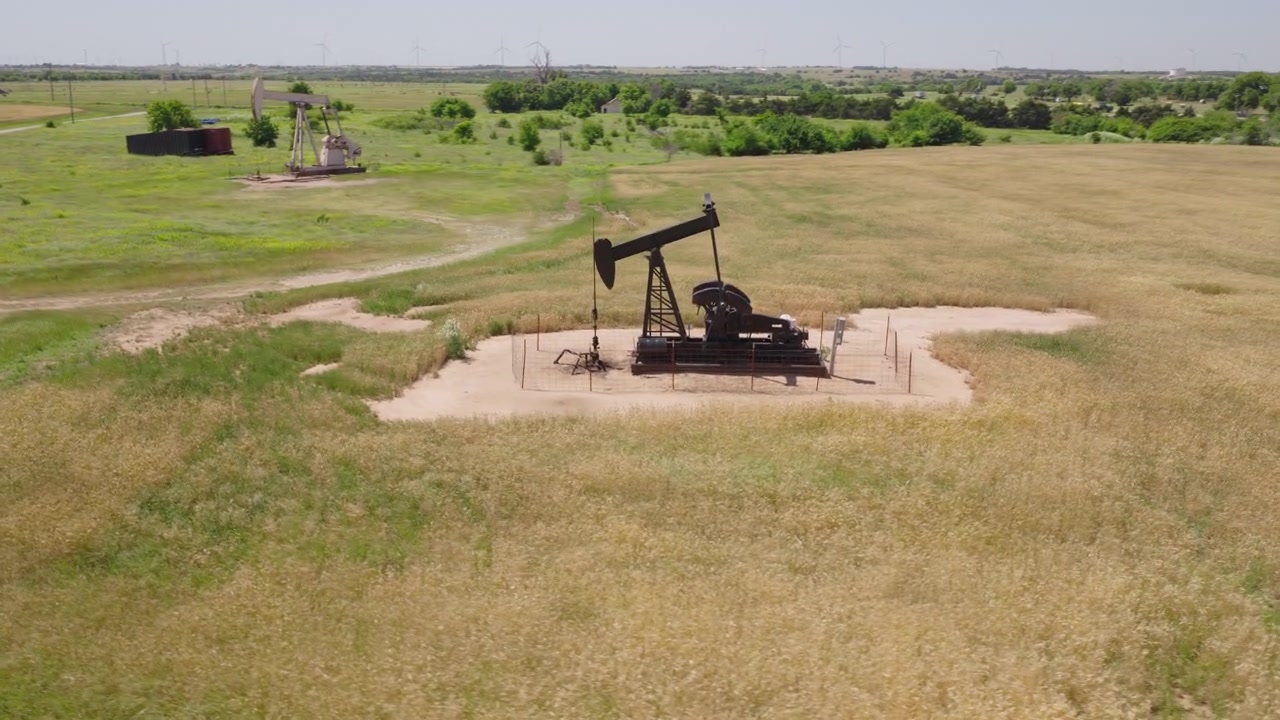 Drone 4K of a wheat field with two oil pump jacks and wind turbines in the background.