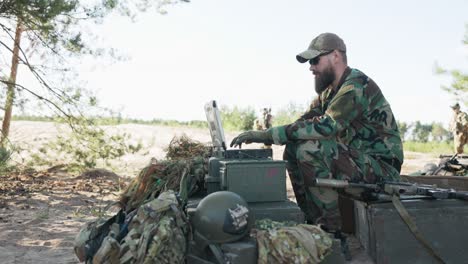 army commander sits behind computer in field and creates reports to be sent to army base man approaches him and shows map they discuss agree on details in background soldiers guarding camp