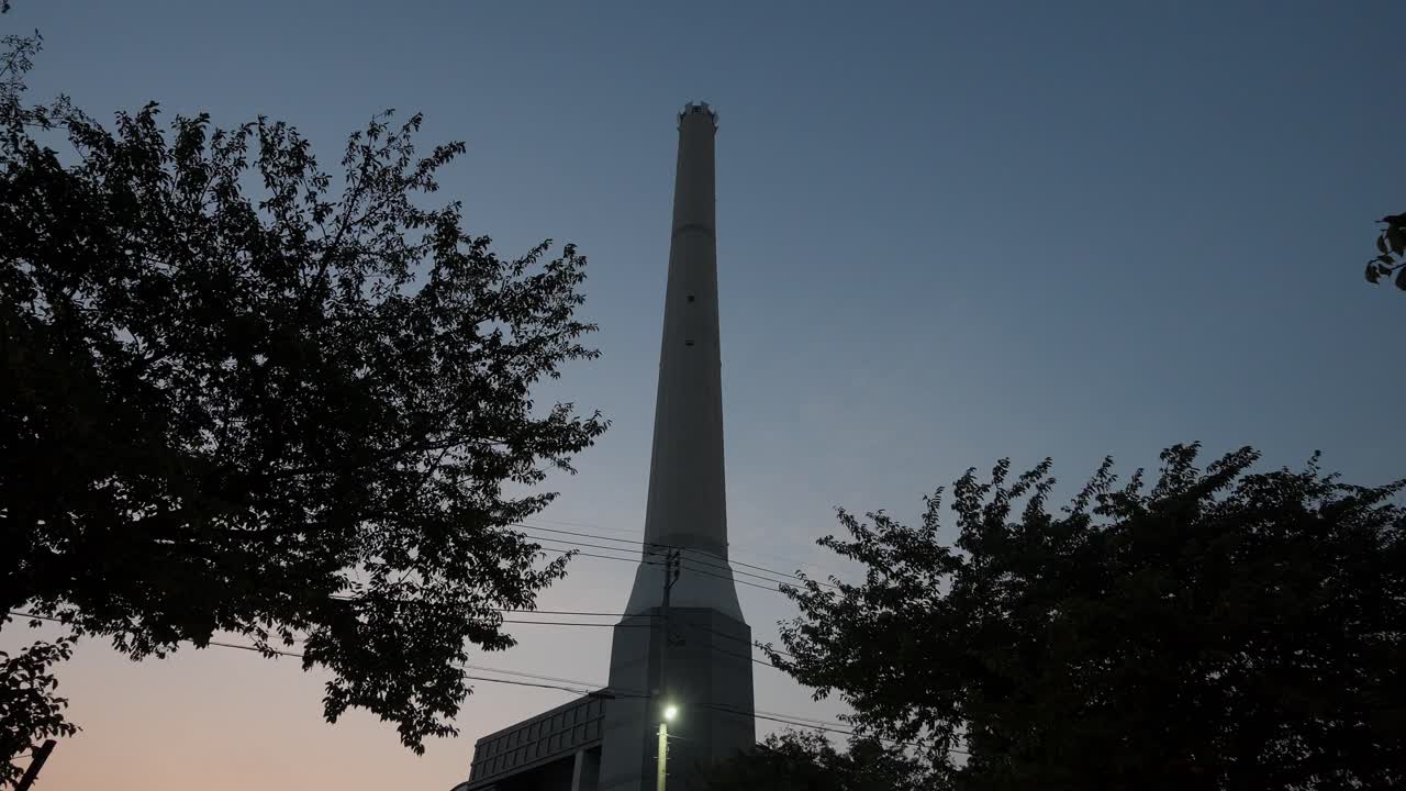 A tall, industrial chimney stands against a twilight sky, surrounded by trees at dusk