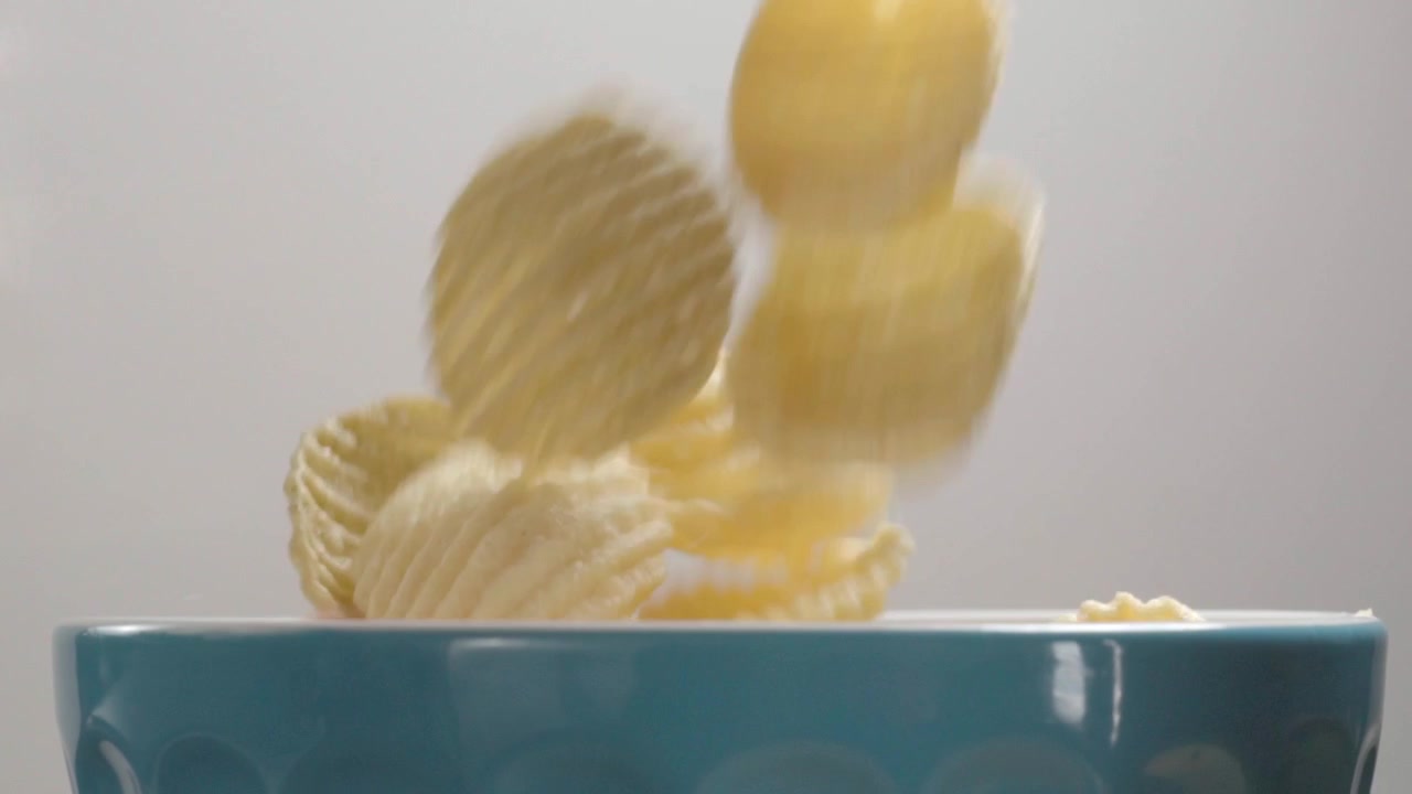Patatina che cade in una ciotola blu, mostrando consistenza croccante e movimento su uno sfondo chiaro e pulito durante la preparazione del cibo