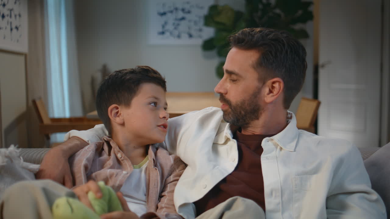 Father embracing son talking evening living room. Cute toddler holding soft toy