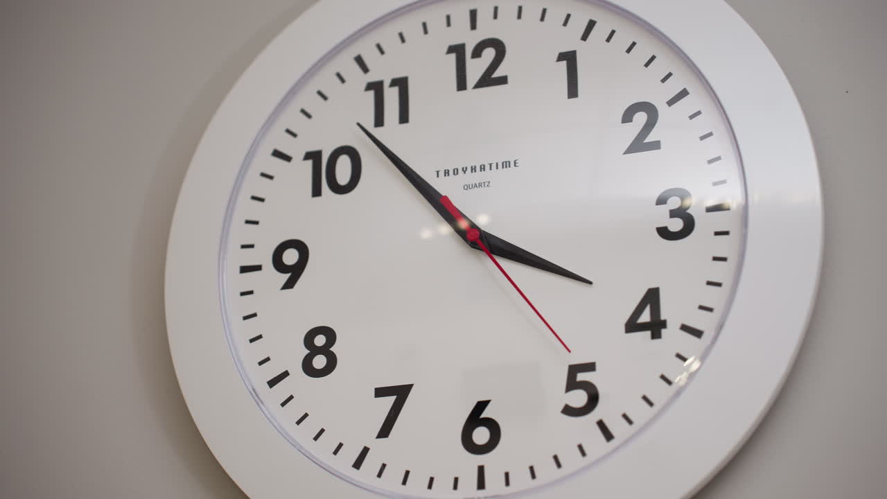 White analog wall clock with bold black numbers and red second hand hanging on light grey wall. Clock shows time as 3:53 with second hand in motion. Modern, clean interior design