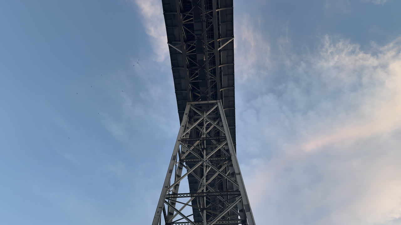 Low-angle shot of the iconic Dom Luís I Bridge in Porto, Portugal, captured from beneath its steel trusses. The towering structure stands out against a pastel-toned sky