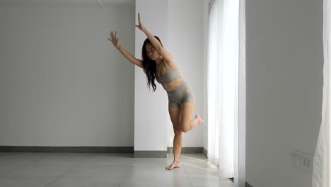 young woman practicing yoga with a standing split pose in a bright room, morning light