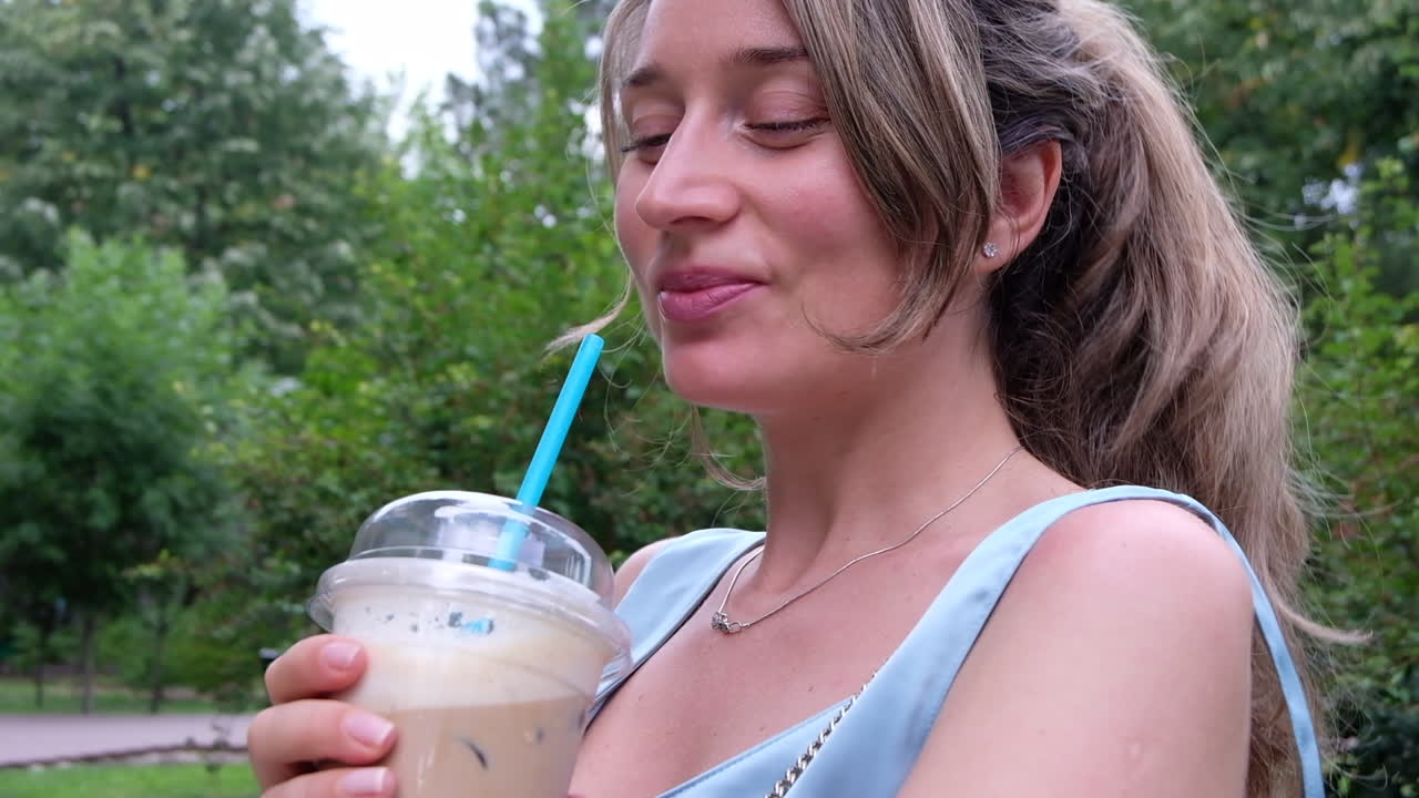 Brunette woman in a blue dress sipping an iced latte in the park
