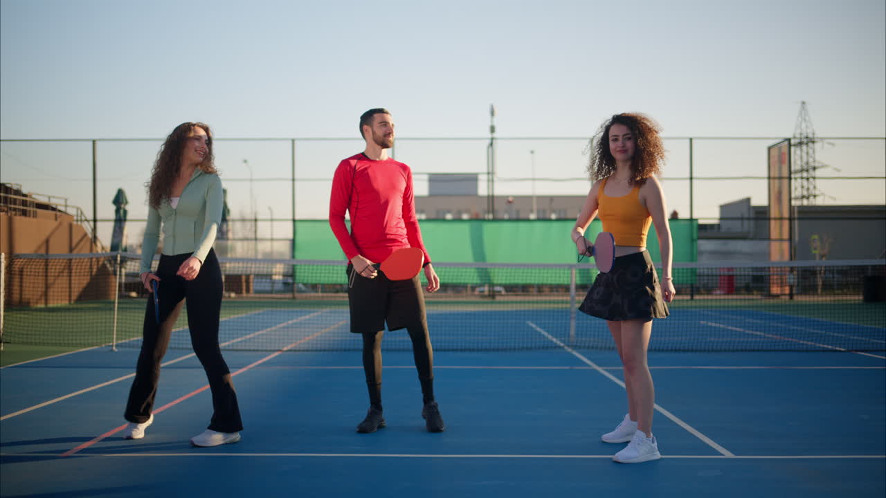 A man in a red shirt teaching two women with curly hair how to play pickleball on a blue court on a sunny day