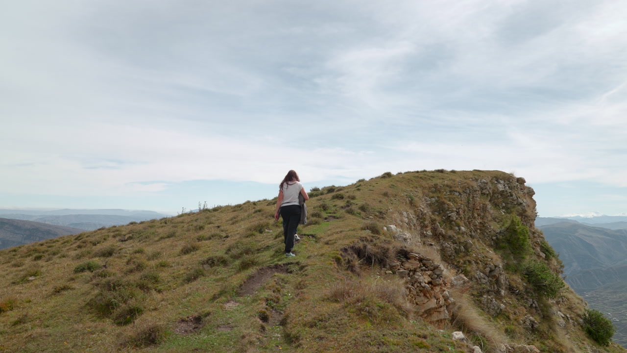Lone Hiker Walking Toward Rocky Edge, Rear Perspective Shows Backpacked Trekker Ascending Ridge, Windblown. Creative