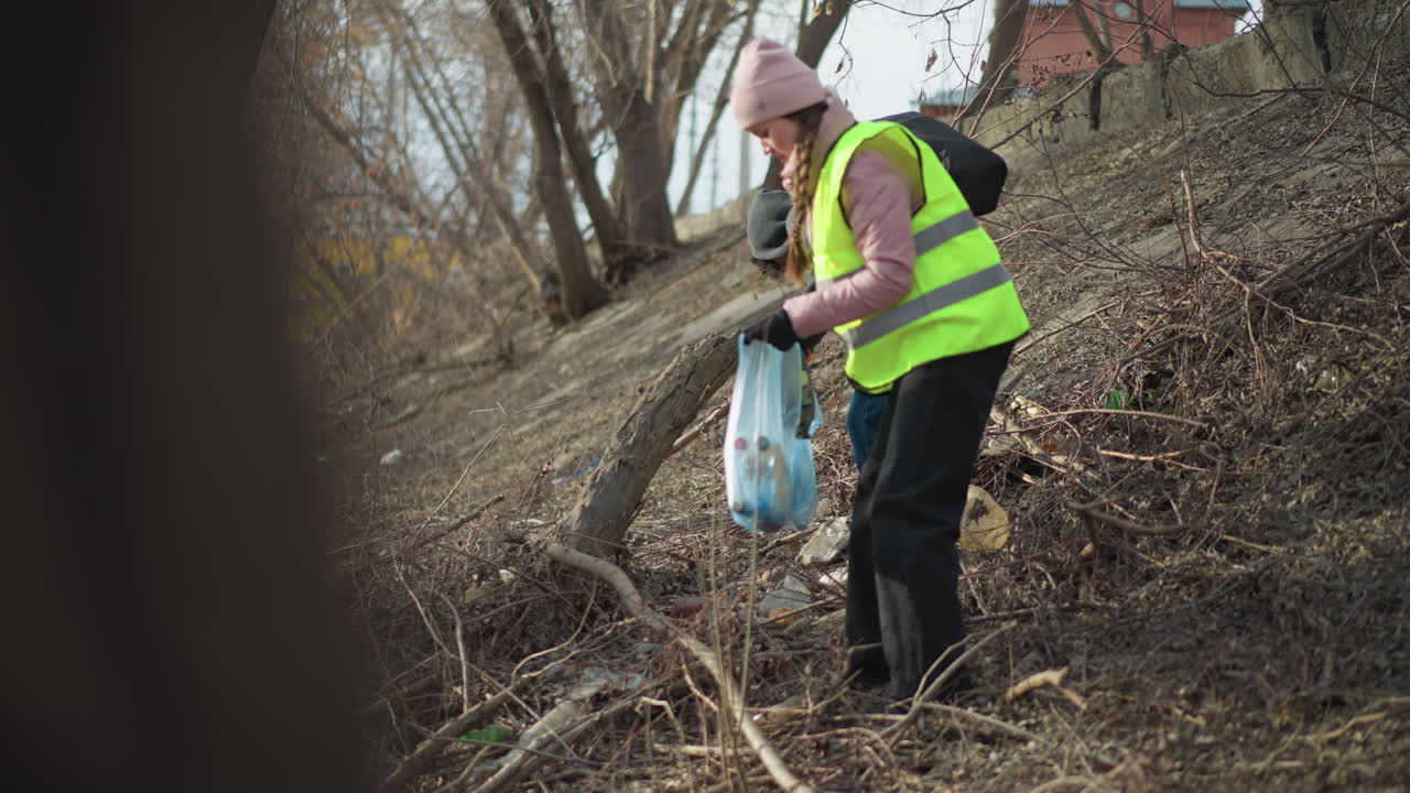 Two volunteers wearing reflective safety vests and gloves collecting trash in blue bag during outdoor cleanup on riverbank, removing litter and debris to protect nature, reduce pollution