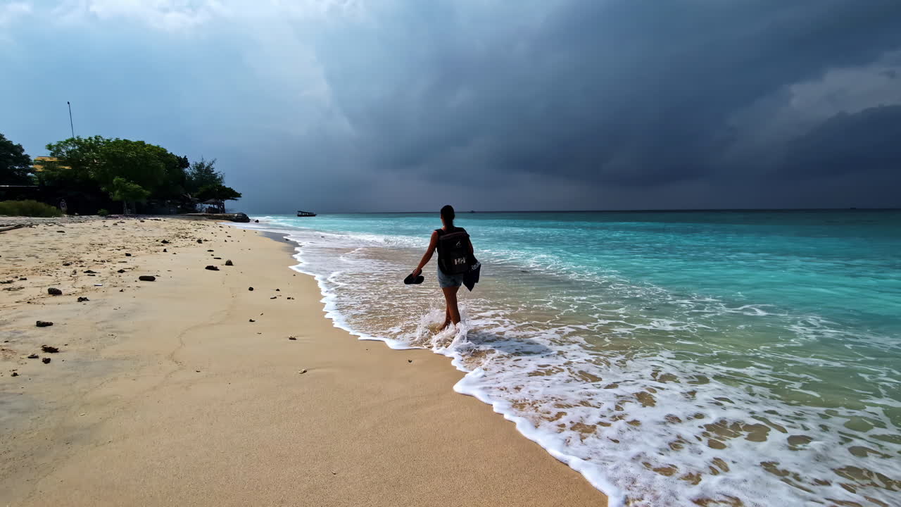 Woman with backpack and barefoot walking along sandy beach on a cloudy day in Indonesia