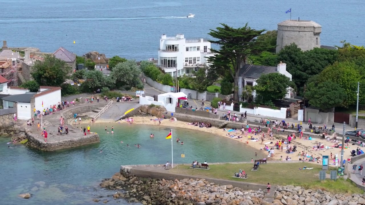 Famiglie nuotano, prendono il sole e si rilassano in una piccola caletta sabbiosa sotto una torre Martello e case sul mare in una luminosa giornata estiva