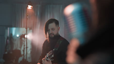 a bearded guitarist playing guitar in a blurry studio room with soft lighting, capturing the musician's focused expression and the blurred microphone in the foreground