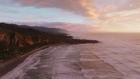 Rising-aerial-view-of-over-Punakaiki-Pancake-Rocks-during-glowing-sunset-over-coastal-landscape,-beach-and-Tasman-Sea-on-the-West-Coast-of-South-Island,-New-Zealand-Aotearoa