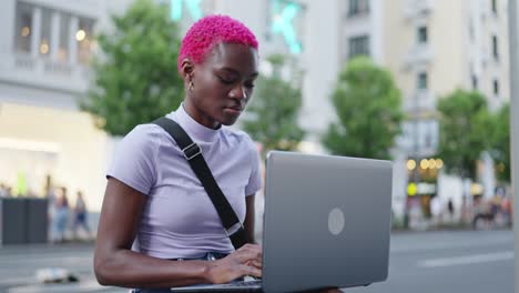 young african american woman with pink hair using laptop outdoors in city