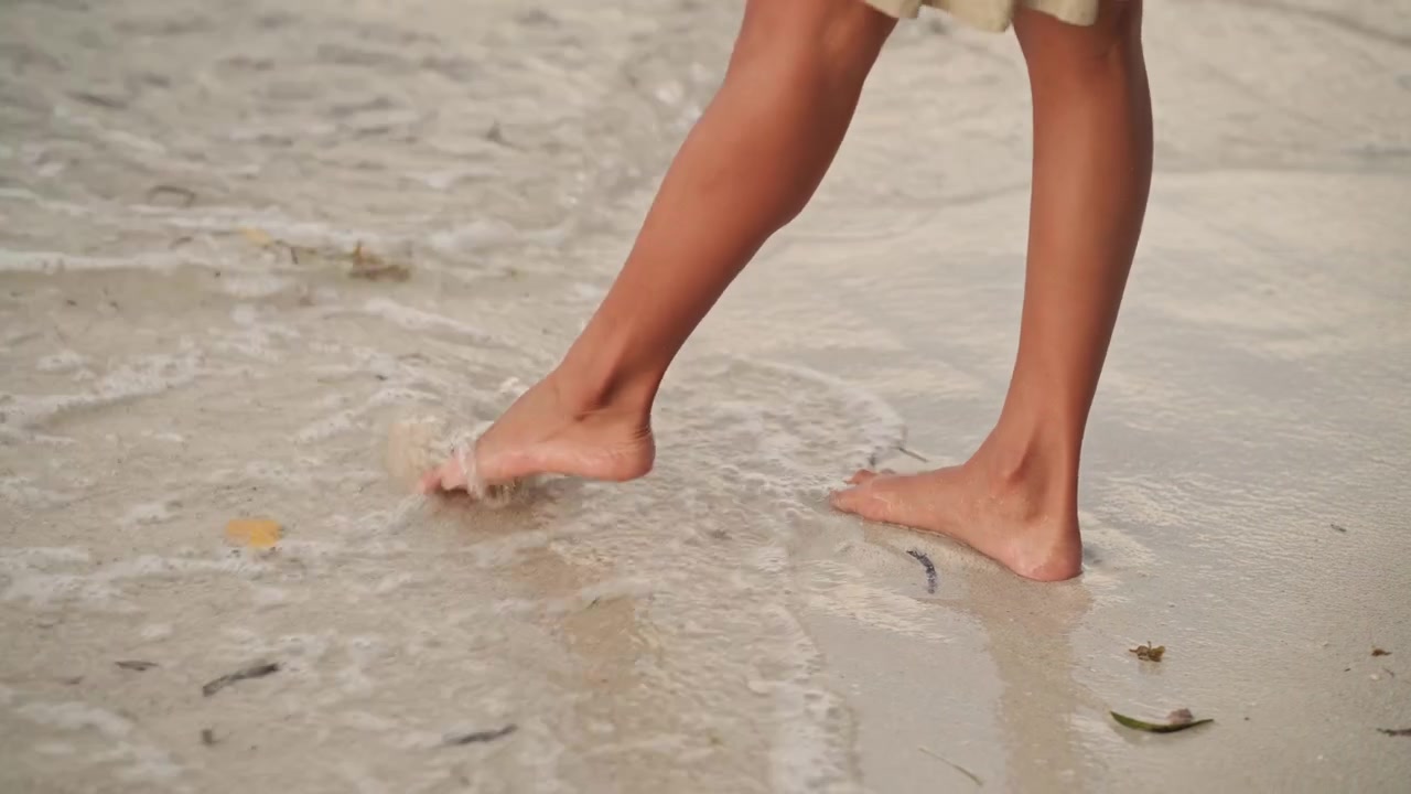 Barefoot woman standing on beach shoreline, toes sinking into wet sand as gentle waves lap at ankles, soft foam and scattered shells, warm golden light, closeup on legs conveying calm and relaxation
