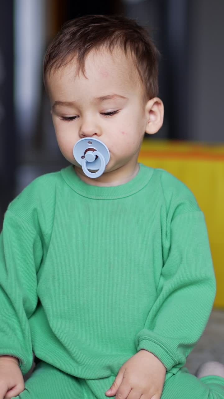 Calm lovely baby sitting on the carpet with toys around. Kid takes a pacifier out of mouth and takes back again. Black cat near the child. Vertical video