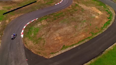 overhead drone view of go-kart racing on a curved asphalt track, captured on a sunny day with clear skies, showcasing two go-karts in motion, contrasting black and white track edges, green grass field