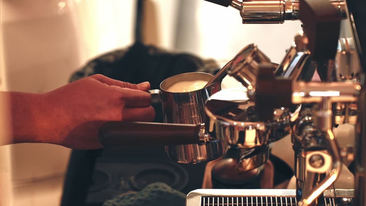 A close-up of a barista's hands holding a metal milk pitcher under the steam wand of an espresso machine