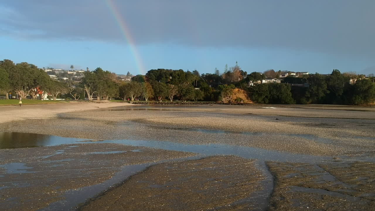 drone volando hacia adelante en una playa a través de la lluvia hacia un arco iris al final de la playa