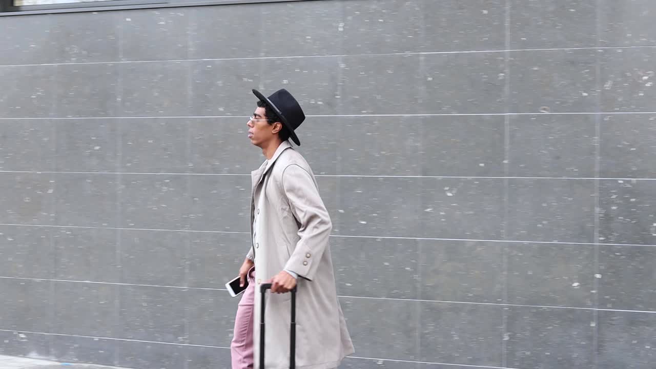 Elegante uomo d'affari etnico con smartphone e bagagli contro un muro di cemento.