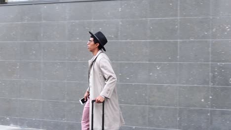 Stylish-ethnic-businessman-with-smartphone-and-luggage-against-concrete-wall