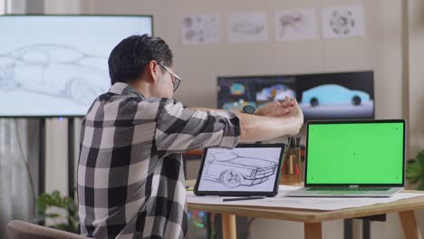 vista lateral de un hombre asiático estirándose mientras dibuja un concepto de coche en una tableta junto a una pantalla verde portátil en el estudio con televisión y computadoras muestra un modelo de coche eléctrico 3d