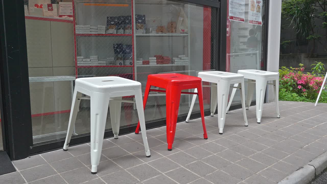 A row of modern metal stools, predominantly white with one striking red stool, are neatly arranged outside a contemporary shopfront.