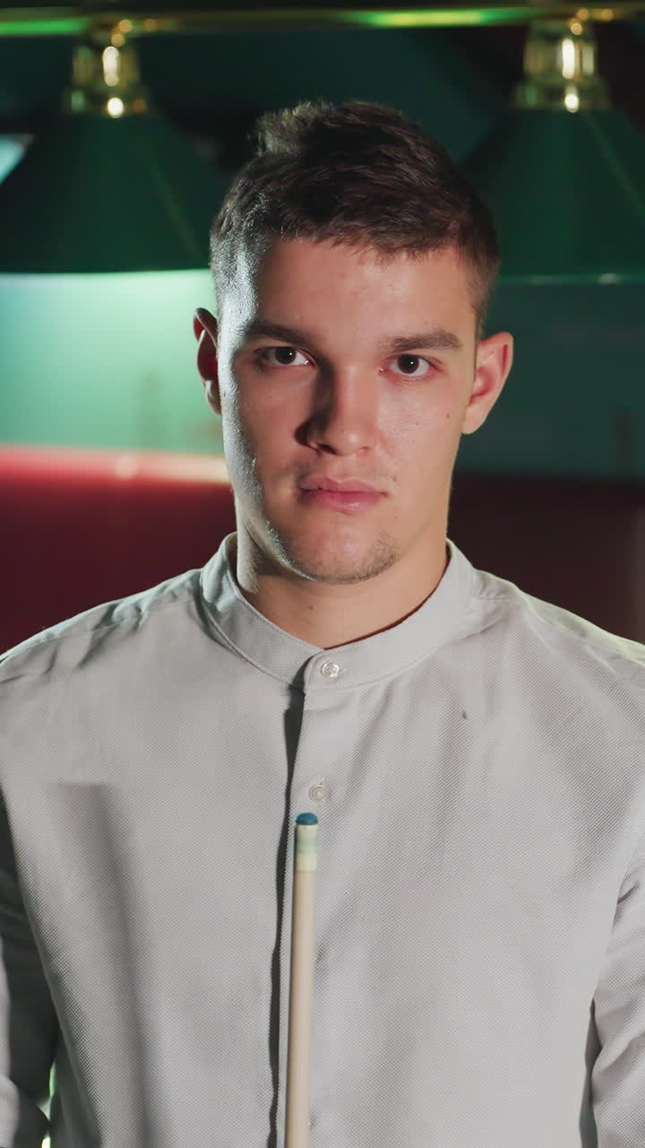 el jugador con cara seria en camisa blanca mantiene la posición enfocada, sosteniendo el palo de la clave en la sala de billar débilmente iluminada. la atmósfera estratégica e intensa destaca la concentración y la preparación para el juego