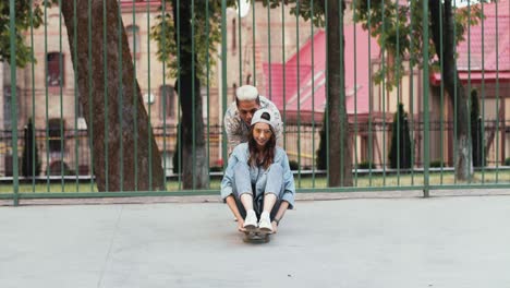 vista completa de una adolescente sentada en una patineta mientras su novio afroamericano la empuja en la calle
