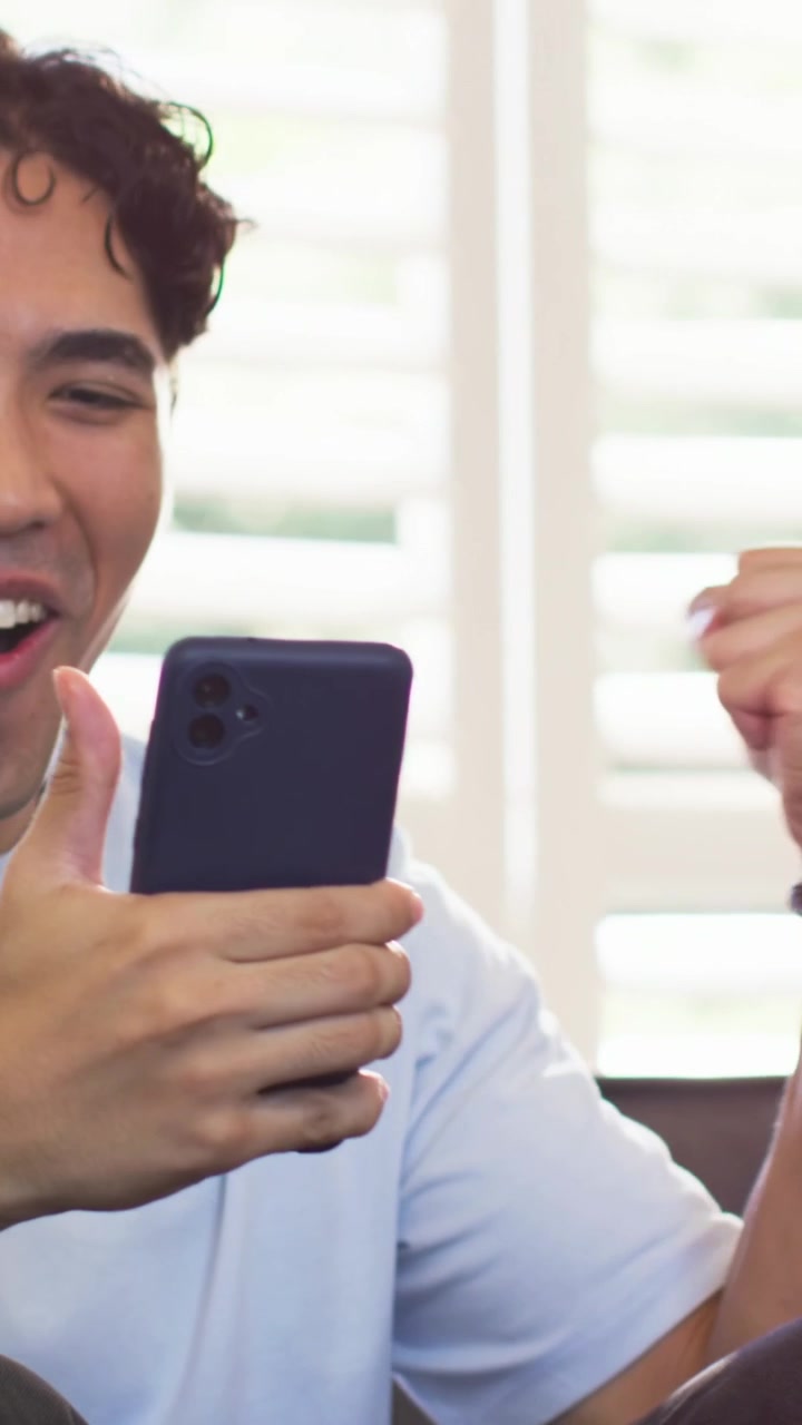 Video verticale: Uomo che ride in camicia blu, tiene il telefono sul divano mostrando un messaggio di vittoria, con spazio per il testo.