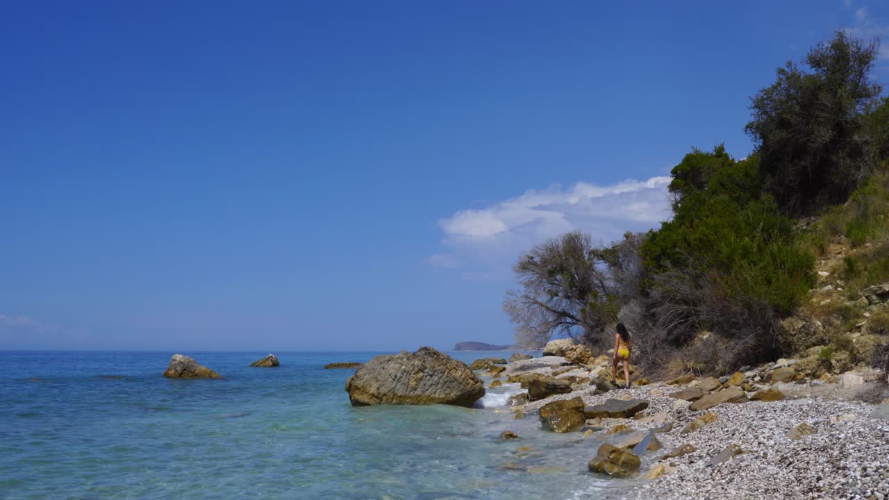 Ragazza in costume da bagno che cammina su una spiaggia rocciosa lungo l'acqua cristallina del mare blu turchese in un'isola greca.