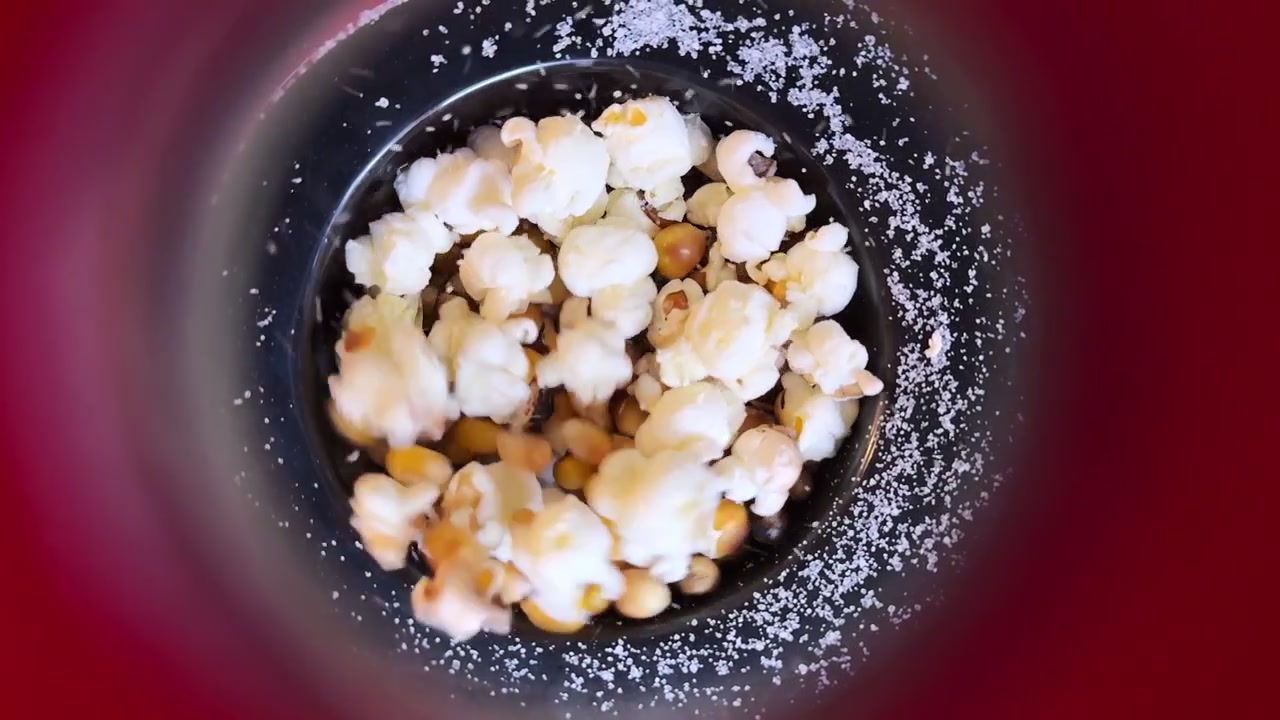 Närbild uppifrån på majskorn som poppar inuti en maskin med saltkorn. Stark inomhusbelysning framhäver övergången från gula frön till vitt fluffigt popcorn under tillredningen.