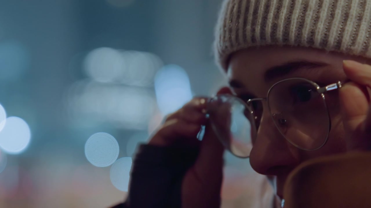 Caucasian student adjusts glasses at night urban bokeh backdrop, reflective eyes, knit beanie, warm coat, cinematic close profile, thoughtful expression, soft light, night city atmosphere