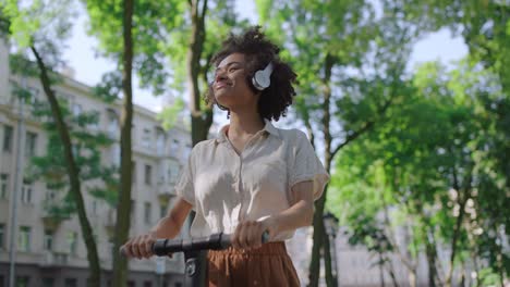happy black woman in headphones riding e-scooter on street, leisure activity
