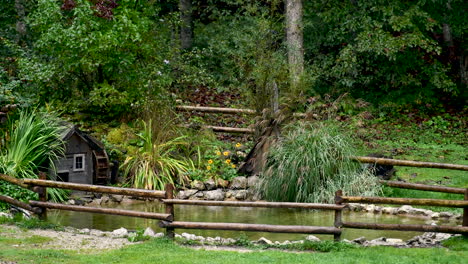 decorative pond with watermill, pond surrounded with flowers and gras, wooden fence built around, steady shot