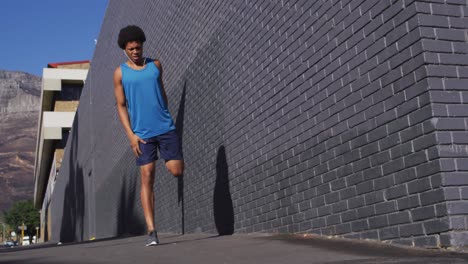 hombre afroamericano en forma haciendo ejercicio en la ciudad, usando auriculares, estirando las piernas en la calle