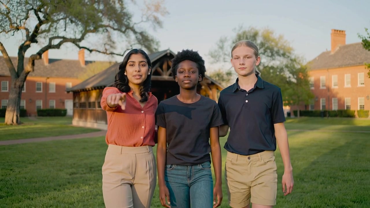 Een wandelend trio op het campusplein bij een paviljoen, waarbij de linker student in een roestkleurige blouse begint te spreken en wijst.