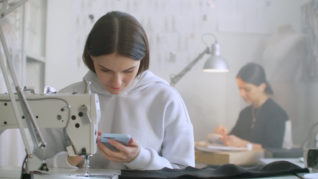 горизонтальное движение камеры. женщина-шивачка смотрит на экран смартфона на рабочем месте. на заднем плане коллега работает и шиет на швейной машине. перерыв в работе шивачек. современная швейная мастерская. интернет-банкинг во время работы
