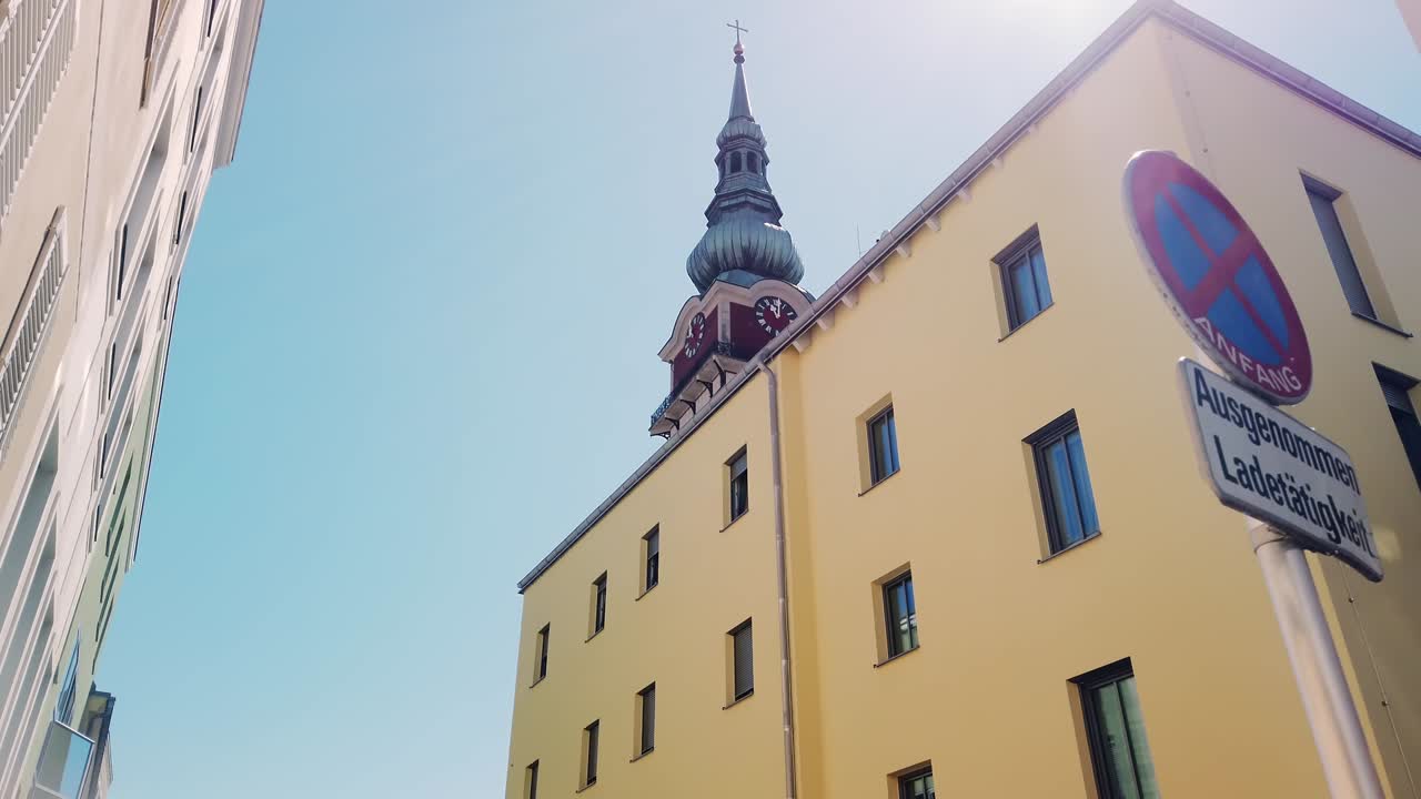 A follow shot captures a yellow building with a tall, ornate spire and clock tower under a clear blue sky, framed by neighboring structures in an urban setting.