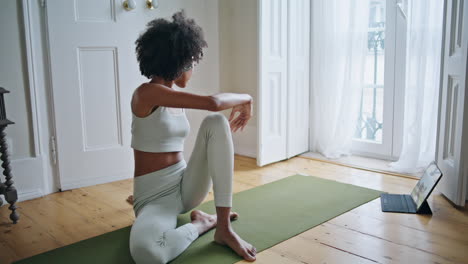 slim girl stretching legs window room. african lady watching tablet yoga lesson