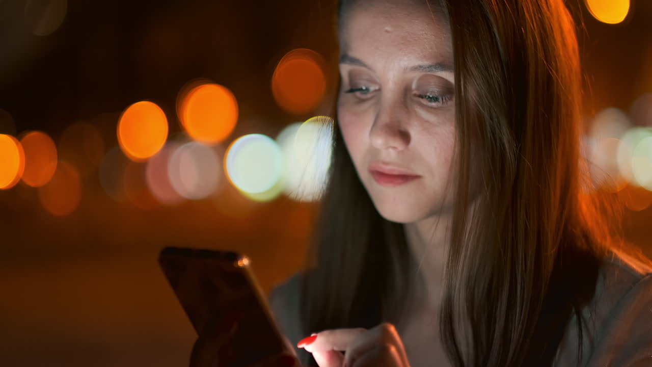 A beautiful young girl in the summer in a big night city keeps a smart phone in shell and writes a message reads e-mail and communicates in chat rooms and social networks keeps a blog looks at the phone screen