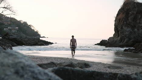 Man-walks-out-of-ocean-on-beach-in-small-secluded-beautiful-cove,-slow-motion