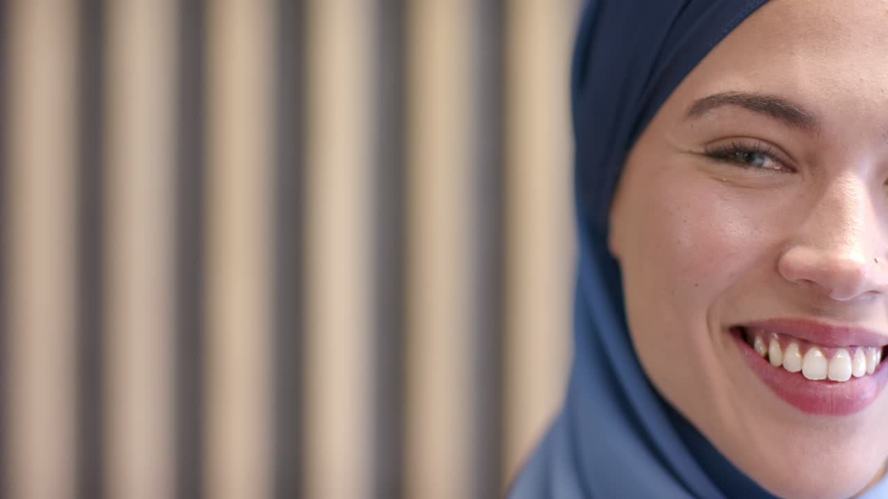 Adult woman in blue hijab reacting to camera, blinking and smiling with studio slats, copy space