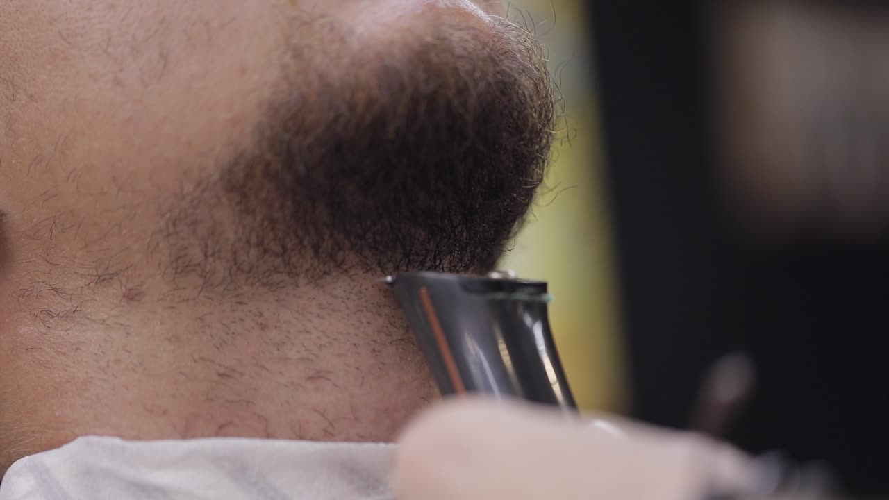 Barber cutting beard using electric trimmer, makes haircut for groom man customer at barber shop