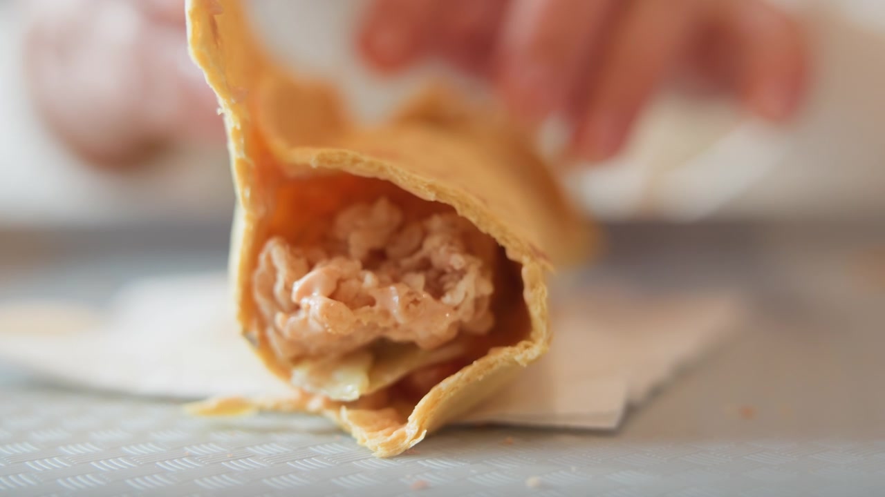 Tampilan makro dari crepe yang disobek terbuka untuk memperlihatkan isian krim parut dengan potongan sayuran kecil, isian tumpah ke serbet sementara jari-jari yang buram memegang subjek dalam cahaya lembut yang hangat.