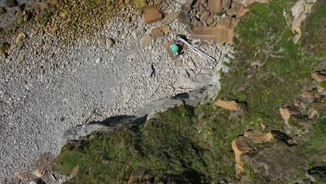 Toma-Superior-En-Un-Acantilado-Por-El-Que-Discurre-Una-Escalera-Que-Baja-A-La-Playa-Pedregosa-Donde-Se-Pueden-Ver-Unas-Sombrillas-Y-Las-Olas-Del-Mar-Rompiendo-Tranquilamente-Contra-Las-Rocas-En-Cantabria,-España.