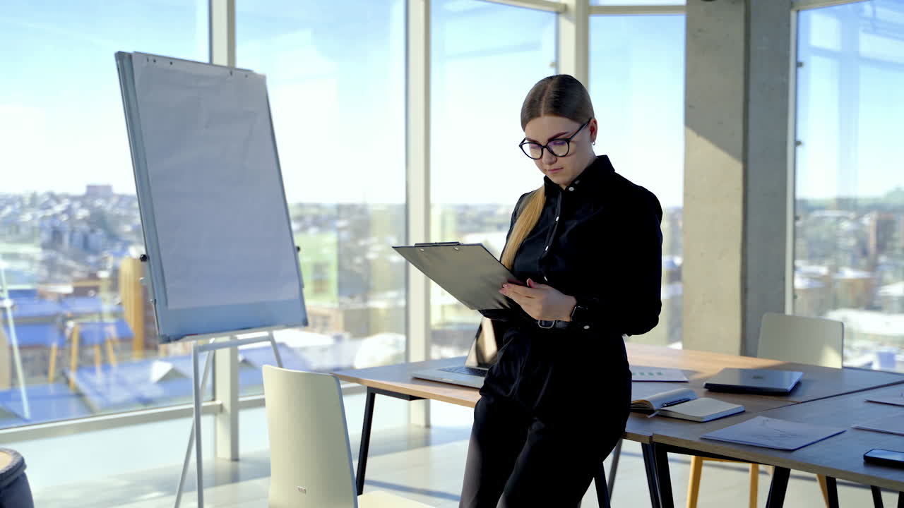 Businesswoman in glasses works in office. Young beautiful lady in elegant black clothes holding a folder and read the information.