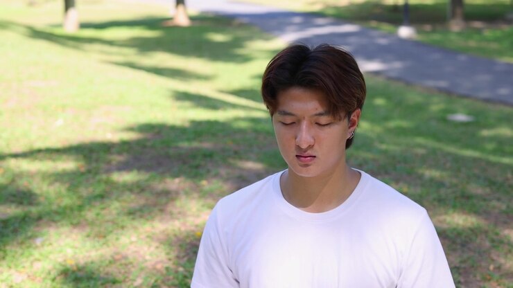 Inquadratura statica media di un giovane uomo asiatico che medita pacificamente in un parco verde e soleggiato.