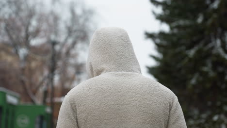 back view of someone in a white winter jacket strolling, blurred background featuring evergreen and bare trees with a distant building, evoking a serene winter scene