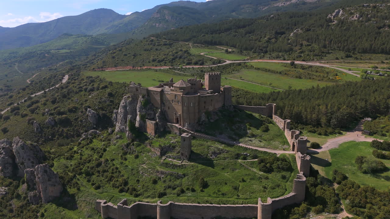Slow orbiting aerial of a medieval castle on a hilltop, featuring fortified stone walls, mountain backdrop and green meadows under clear skies. Scenic and historical rural landscape